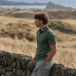 Model wearing the green Deep Cypress Biome polo and gray trousers beside a coastal stone wall.
