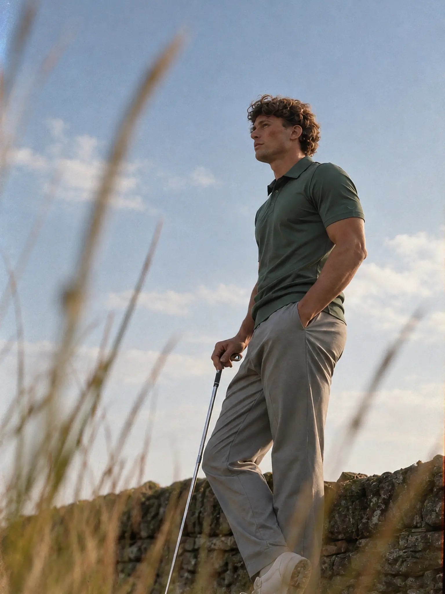 Full-length side view of the Deep Cypress Biome polo worn with gray trousers and a golf club.
