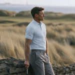 Model wearing the light blue Glacier Biome polo and gray trousers beside a coastal stone wall.