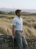 Model wearing the light blue Glacier Biome polo and gray trousers beside a coastal stone wall.