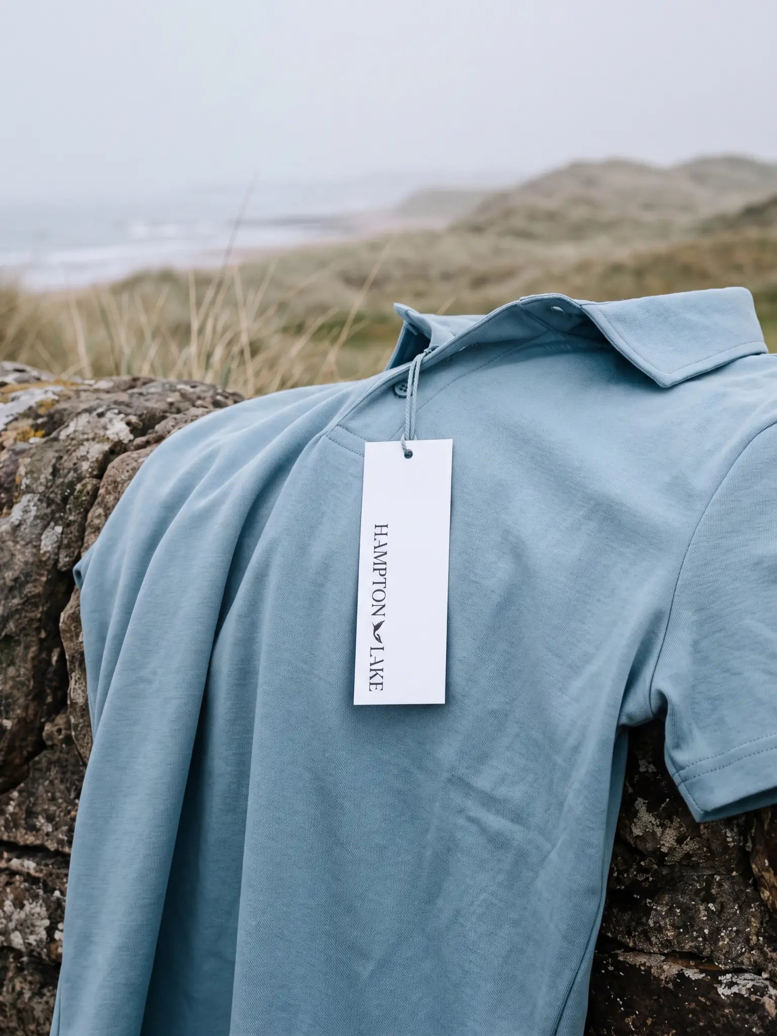 Light blue Glacier Biome polo laid on a stone wall with a Hampton & Lake hangtag.