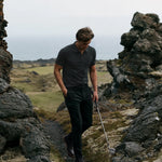 Model wearing the charcoal Volcanic Ash Biome golf polo while walking between coastal rocks with a golf club.