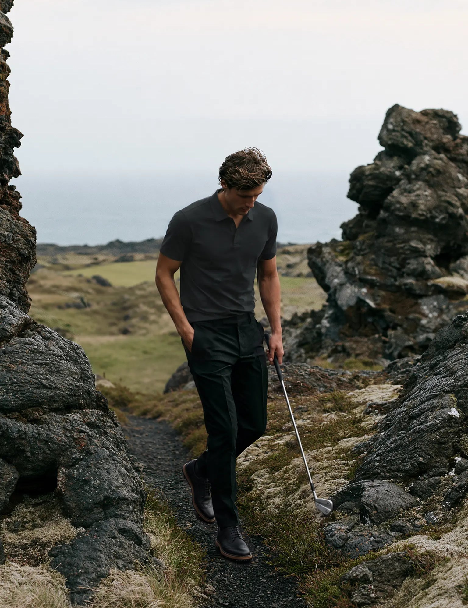Model wearing the charcoal Volcanic Ash Biome golf polo while walking between coastal rocks with a golf club.