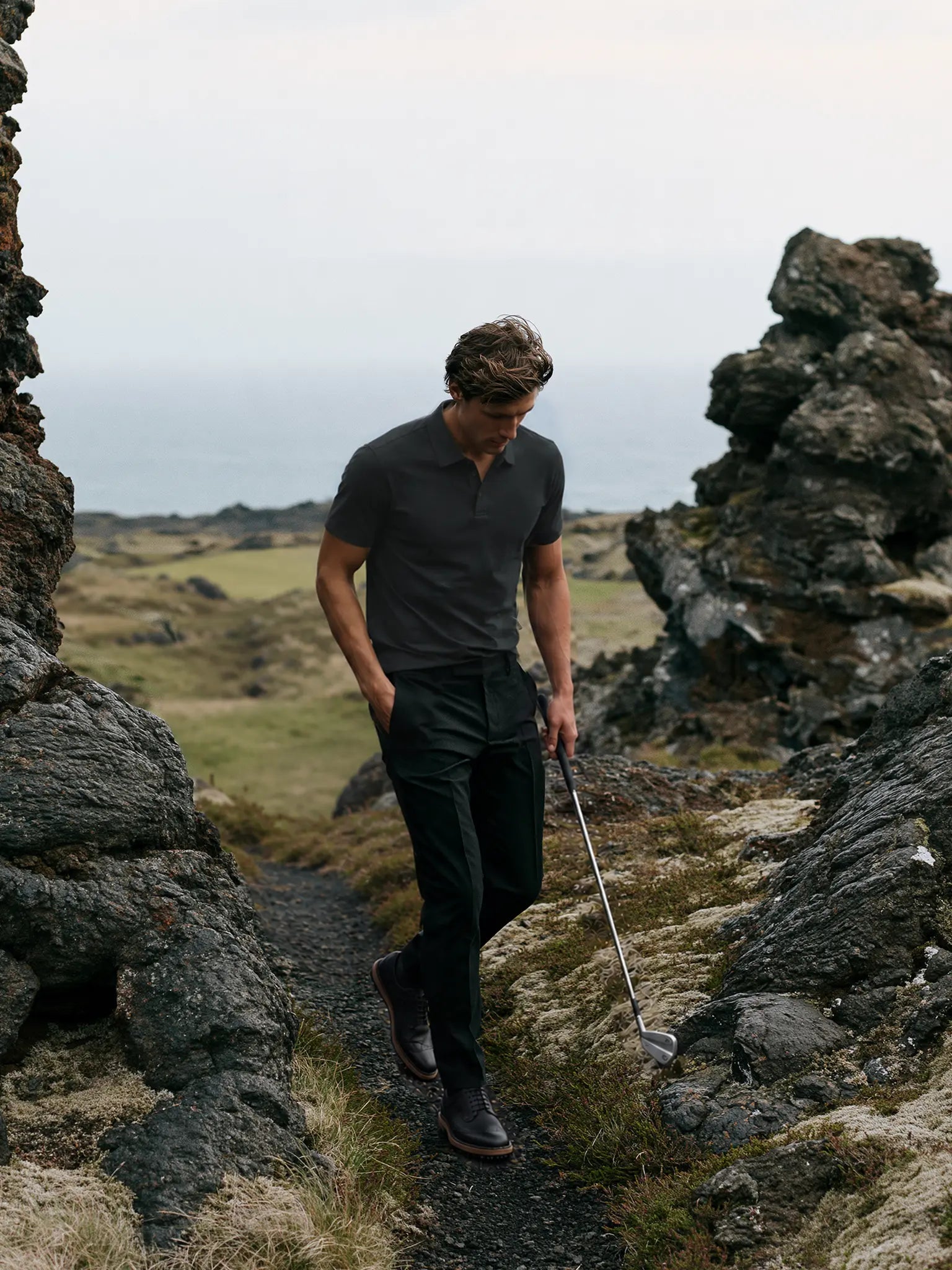 Model wearing the charcoal Volcanic Ash Biome golf polo while walking between coastal rocks with a golf club.