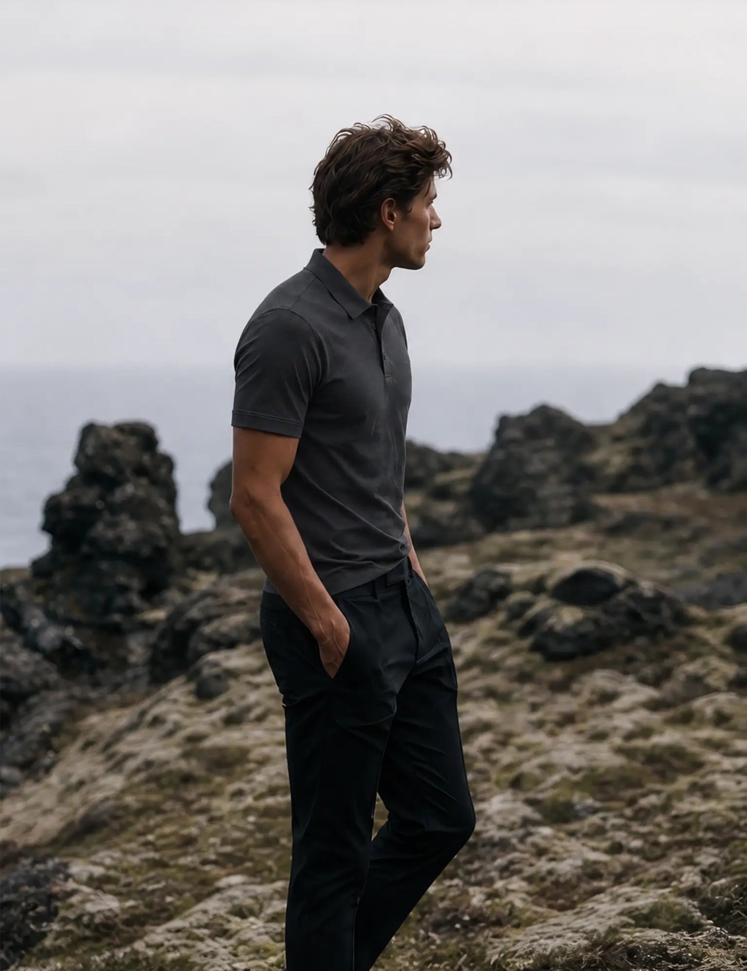 Side view of the Volcanic Ash Biome polo worn with black trousers beside a rocky coastline.