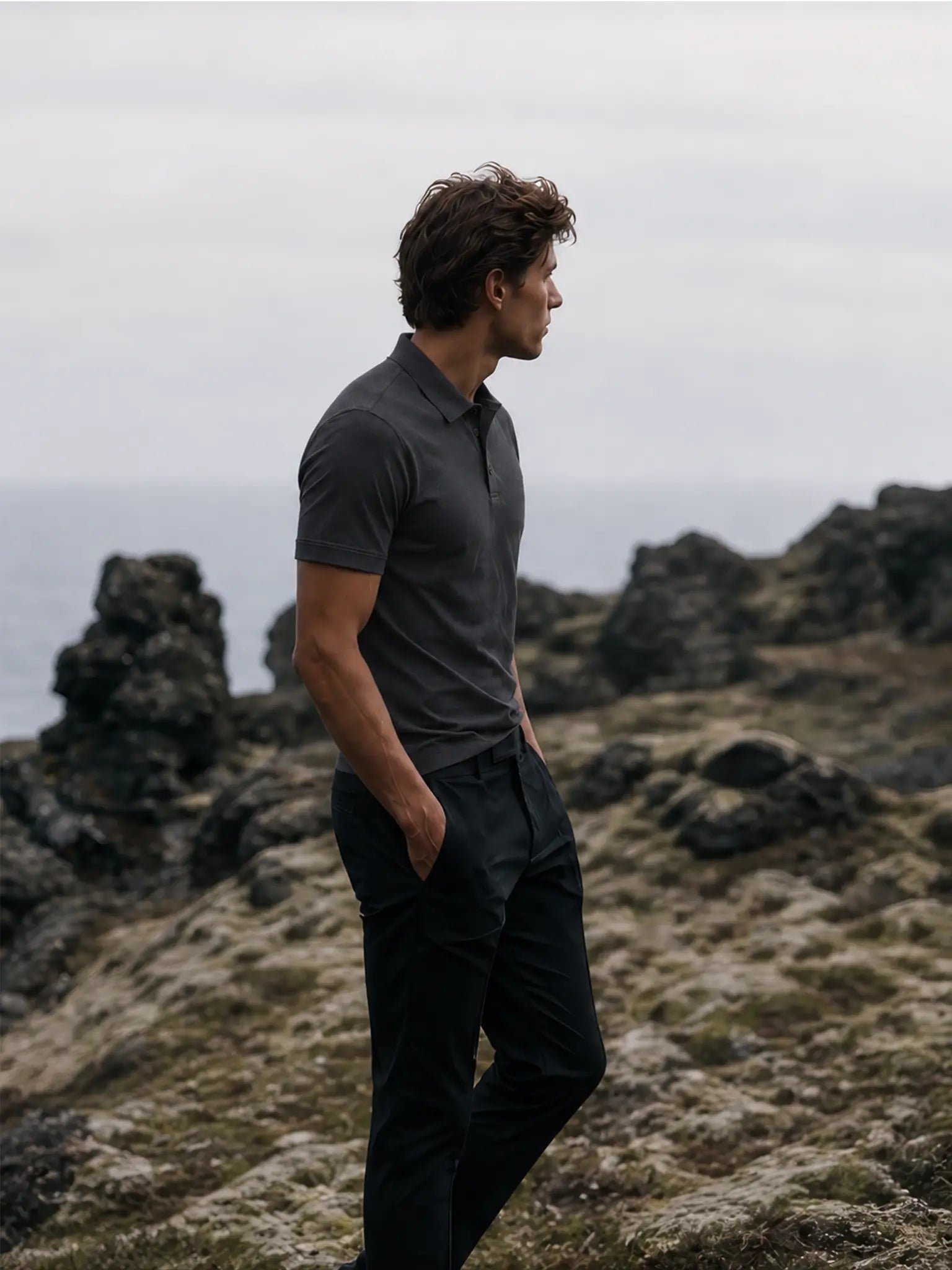 Side view of the Volcanic Ash Biome polo worn with black trousers beside a rocky coastline.