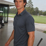 Front three-quarter view of a model wearing the Volcanic Ash Biome polo beside a golf green.