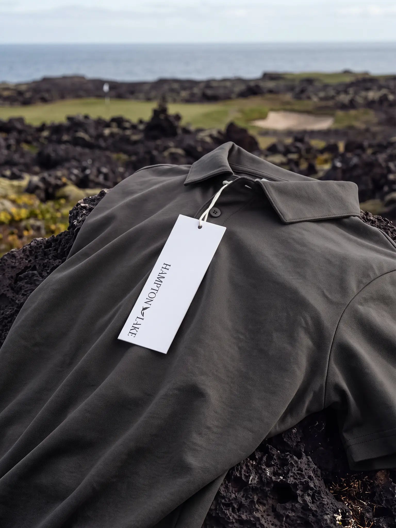 Charcoal Biome polo laid across coastal rocks with a Hampton & Lake hangtag.