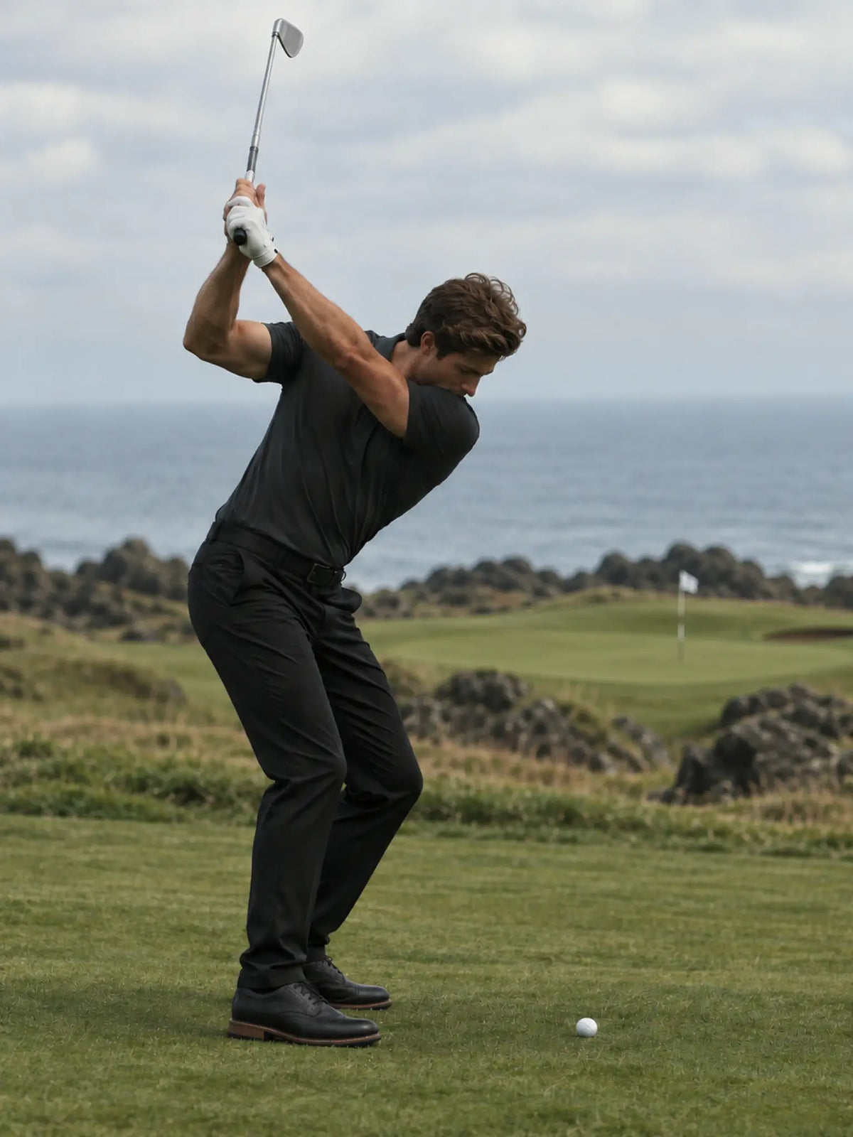 Golfer at the top of a backswing wearing the charcoal Volcanic Ash Biome polo on a coastal course.